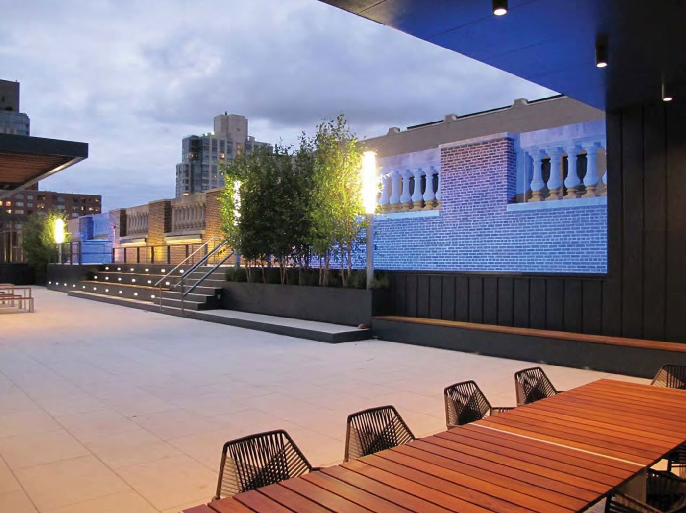 The terrace at dusk with steps, planters, a lit brick parapet and a long timber table.