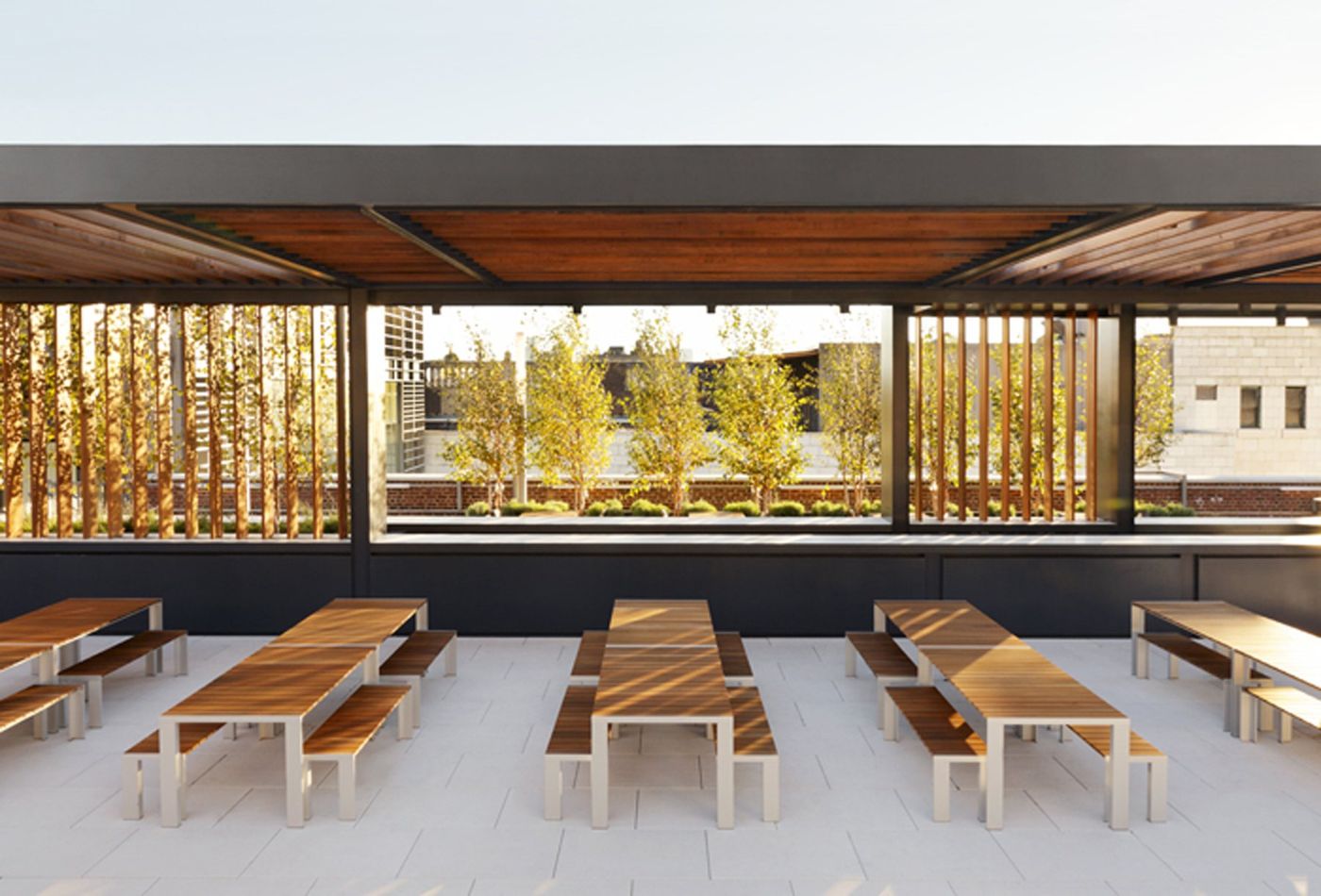 Rows of timber tables and benches beneath the pavilion roof, planting behind.