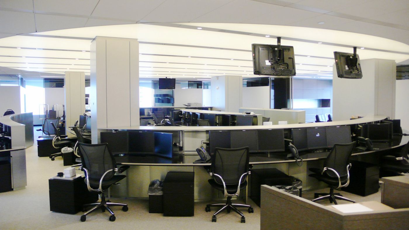 Curved trading desks with screens under a luminous ceiling, columns between the rows.