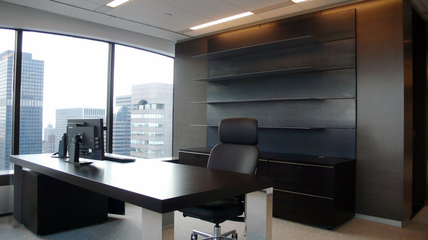 A desk set in front of a dark timber wall of shelves and cabinets, windows to the left.