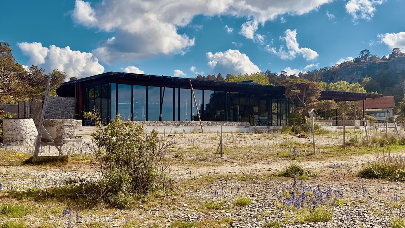 A long glazed hall on a black steel frame beyond dry meadow and shrubs.
