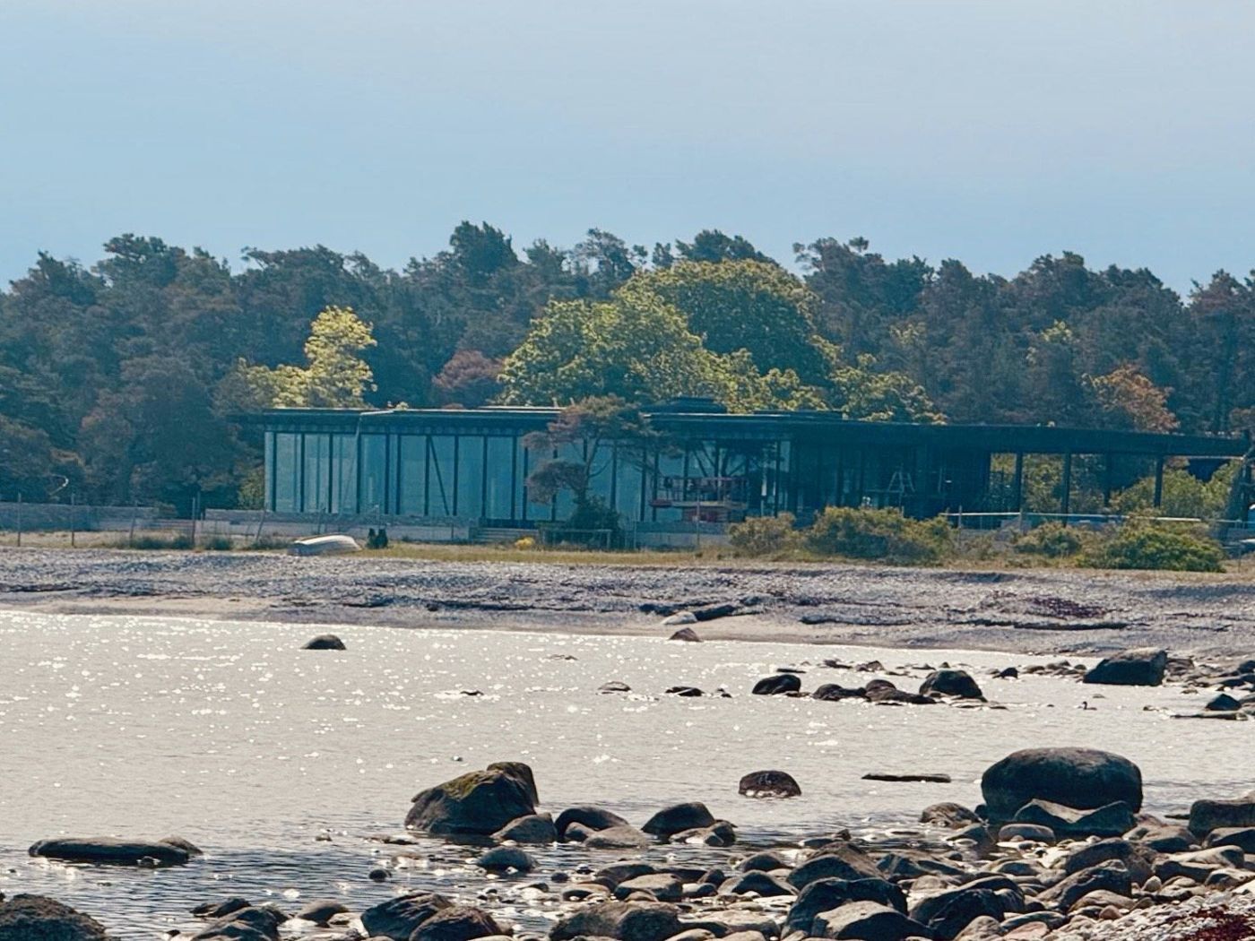 The low building at the tree line seen across a stony shore and calm water.