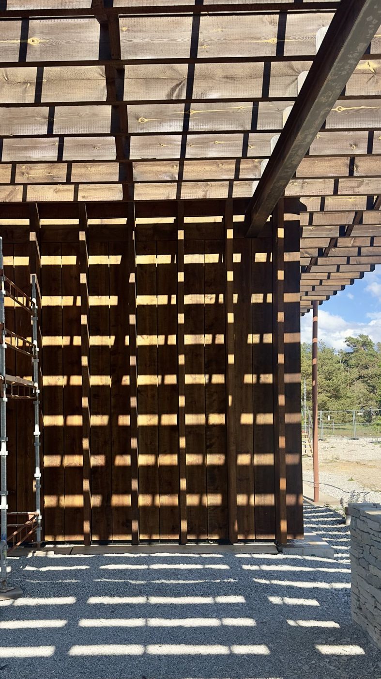 Sunlight through a timber screen and roof slats casts a grid of shadows on the ground.