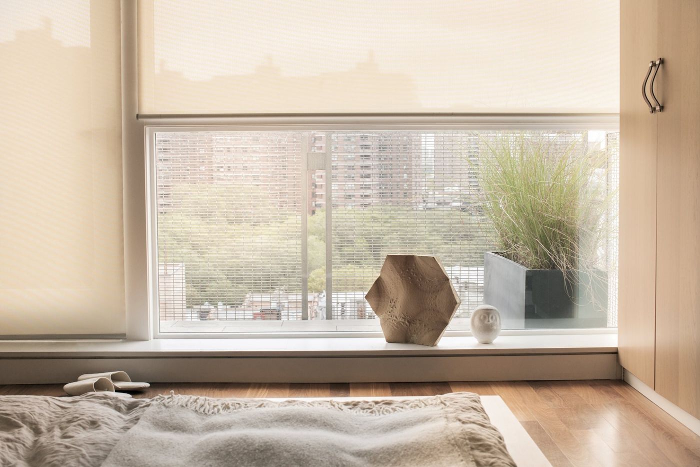A low bed before a wide window with a blind, trees and buildings outside.