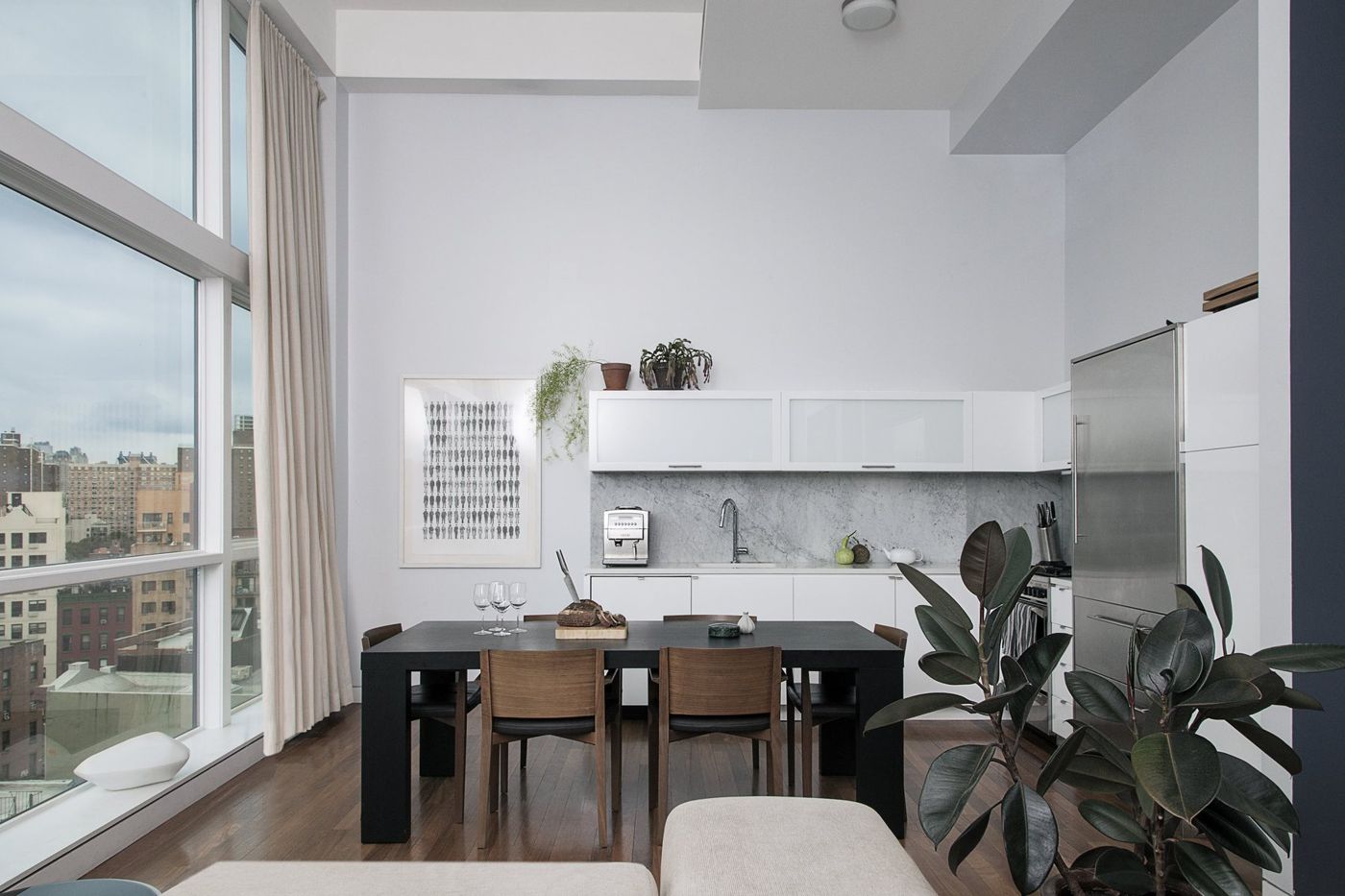 A dining table and chairs before a white kitchen, tall windows and a city view to the left.