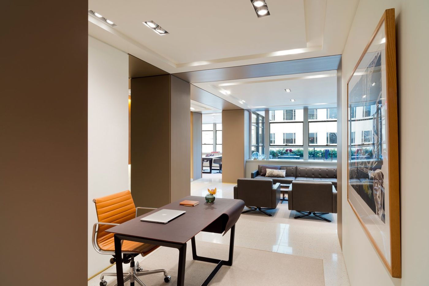 A dark desk and orange chair in front of the lounge, windows and a framed print beyond.