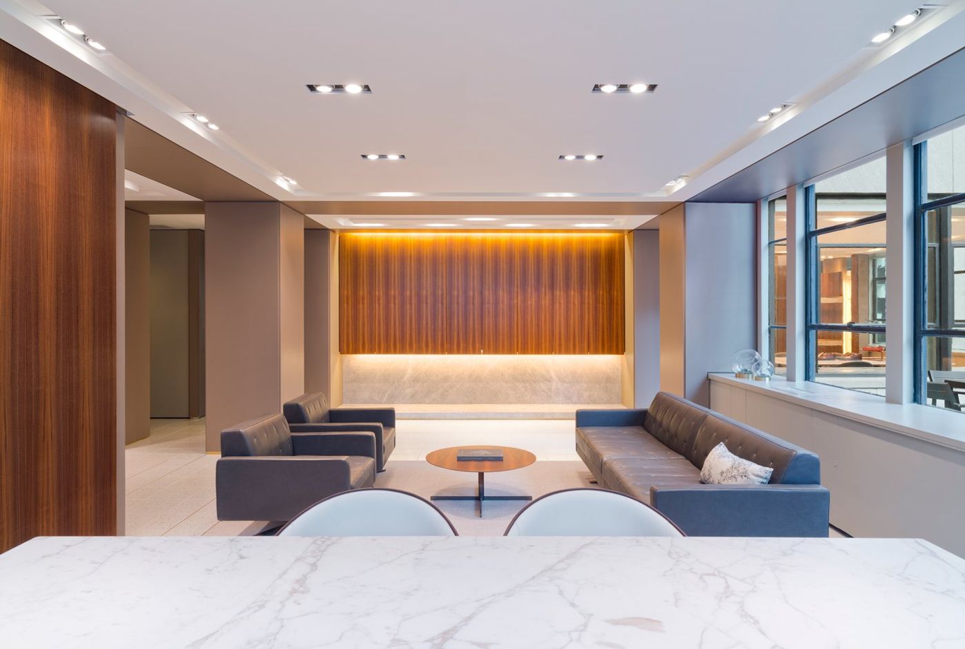 Two sofas face each other before a lit wall of walnut panels, a marble table in front.