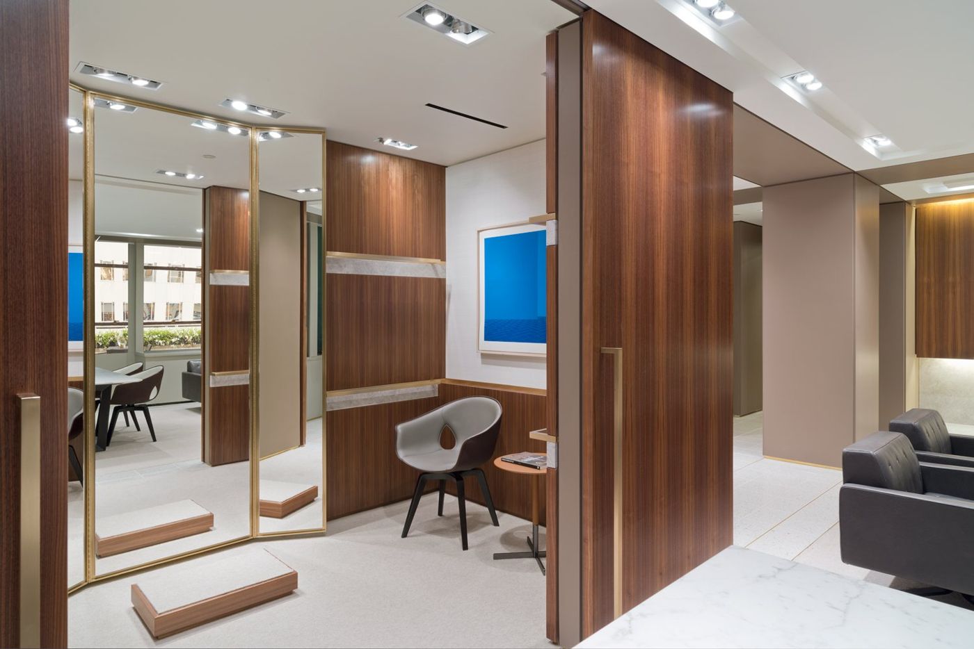 A tall brass-framed mirror and walnut panels in a fitting room, a blue painting on the wall.