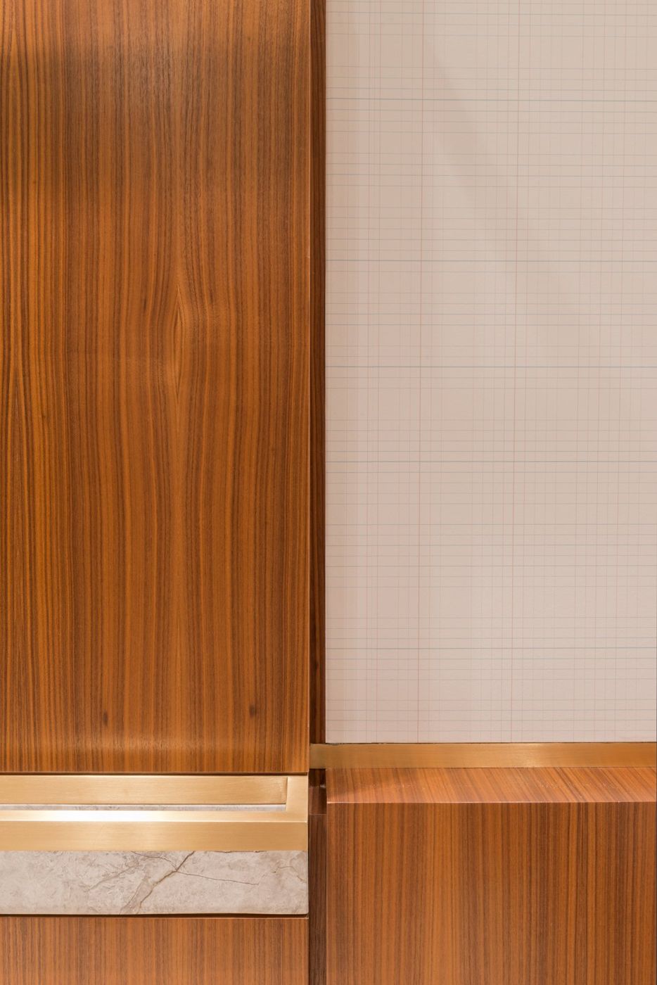 Close view of a walnut panel meeting a brass and stone rail against a gridded wall covering.